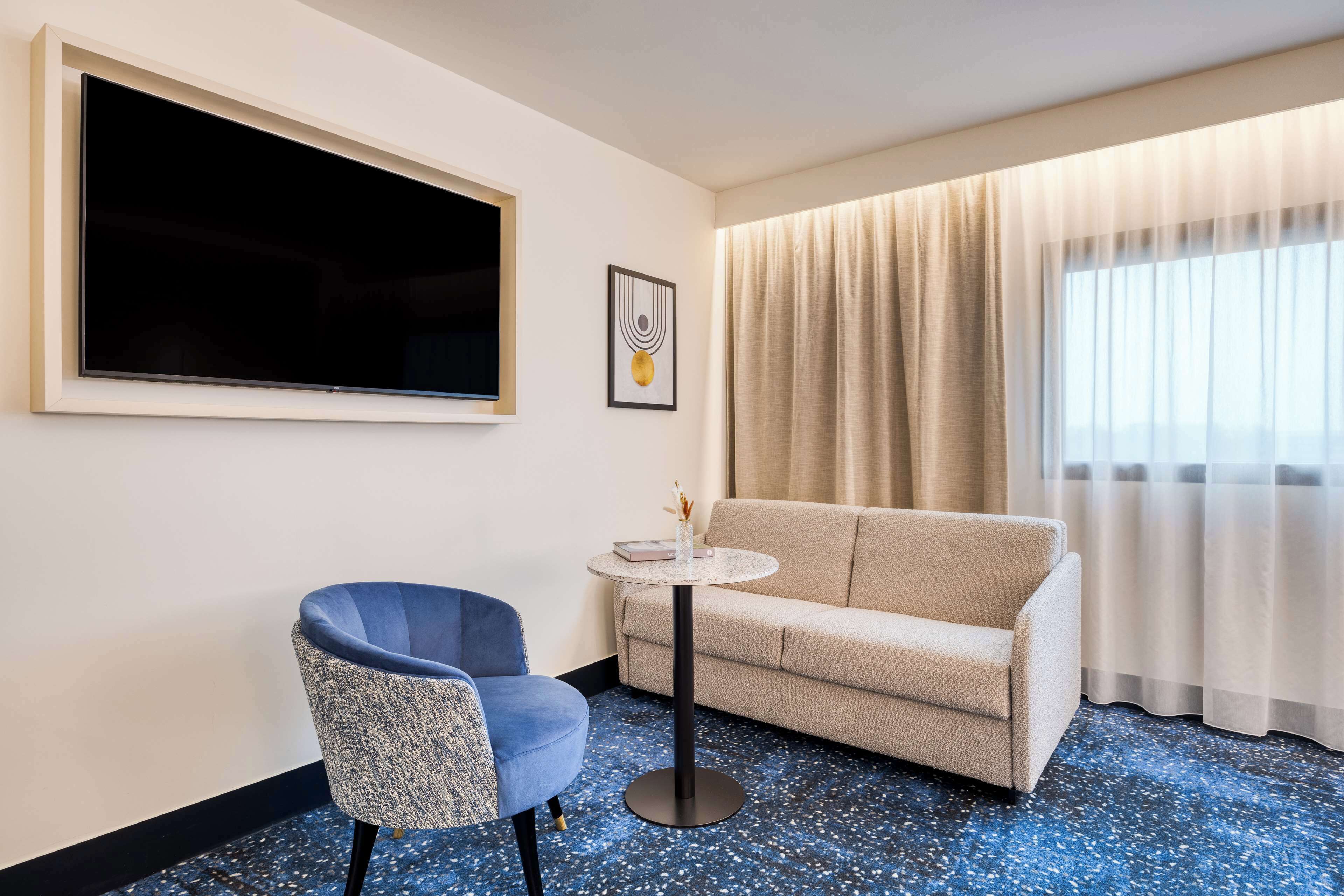 A hotel room with a sofa, a chair, a small round table, a flat-screen TV, and a large window with curtains.