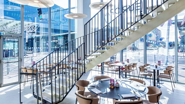 A modern indoor space with a staircase, round tables, and chairs, large windows, and ceiling lights.