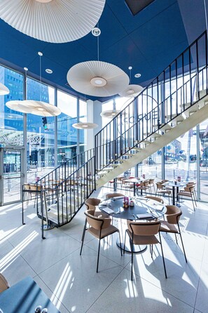 Un espacio interior moderno con una escalera, mesas redondas, sillas, ventanas amplias e iluminación en el techo.