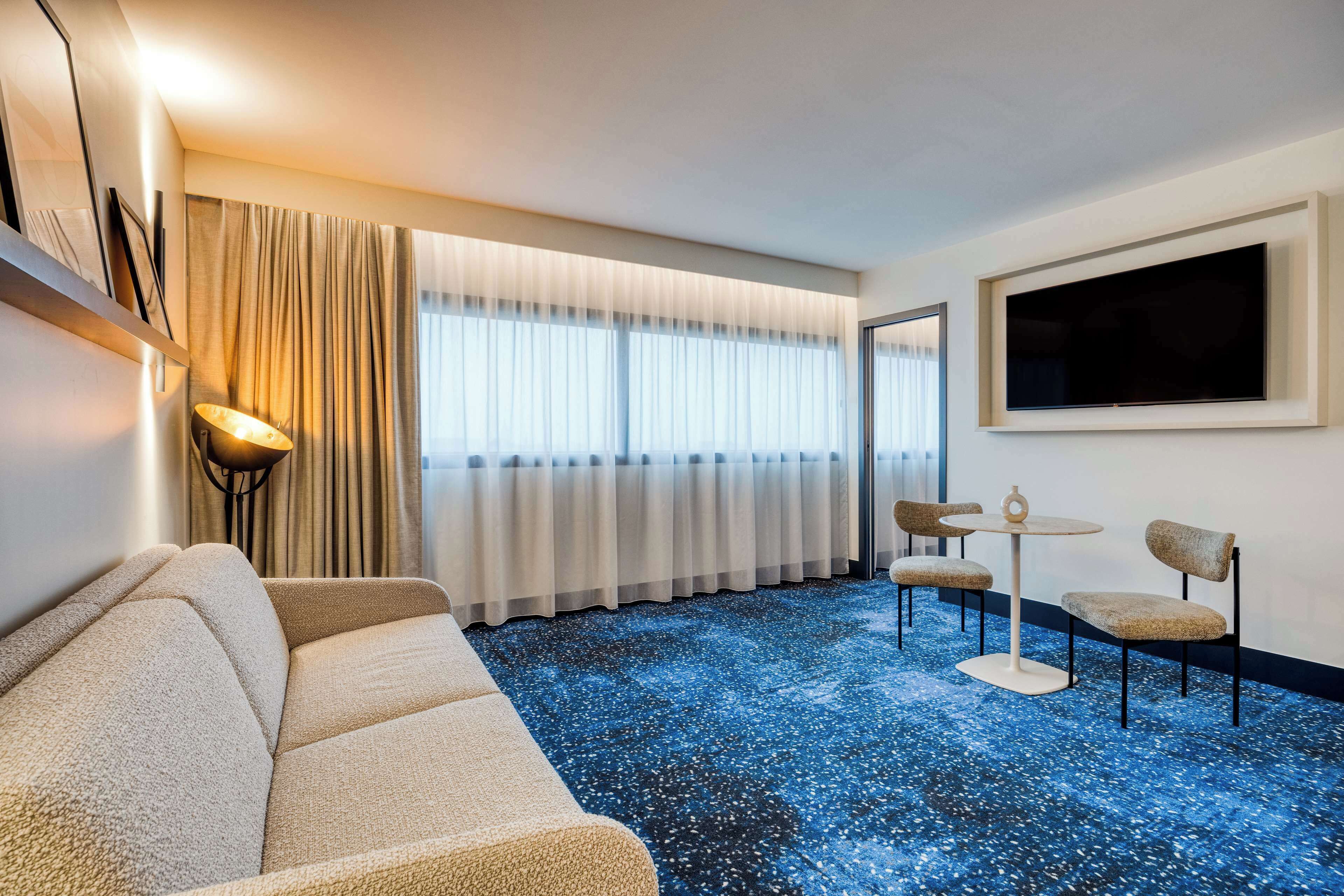 Ein Hotelzimmer mit Sofa, zwei Sessel, einem kleinen Tisch, einem Fernseher und einem großen Fenster mit Vorhängen.