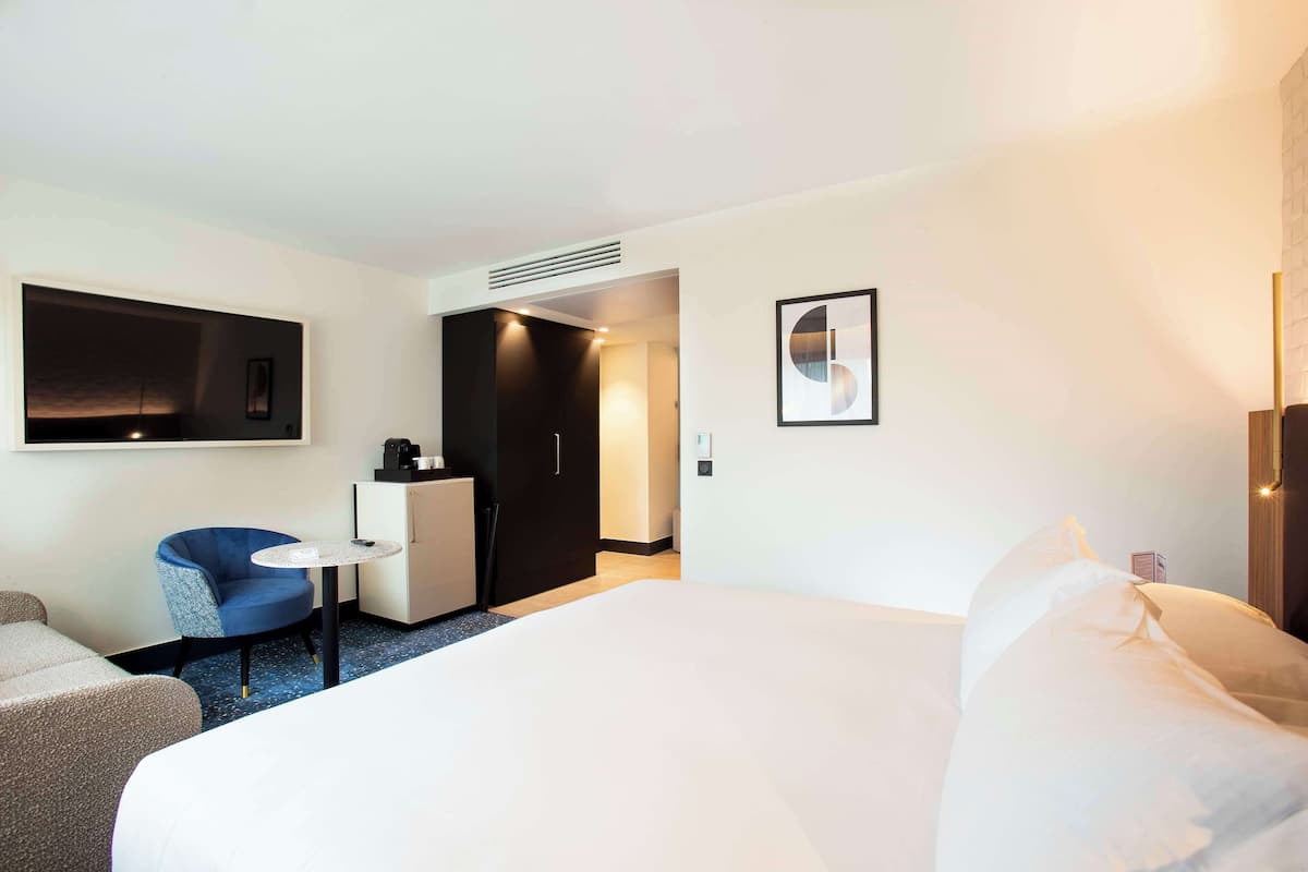 Une chambre d’hôtel avec un grand lit, une petite table ronde, une chaise bleue, un bureau et une armoire.