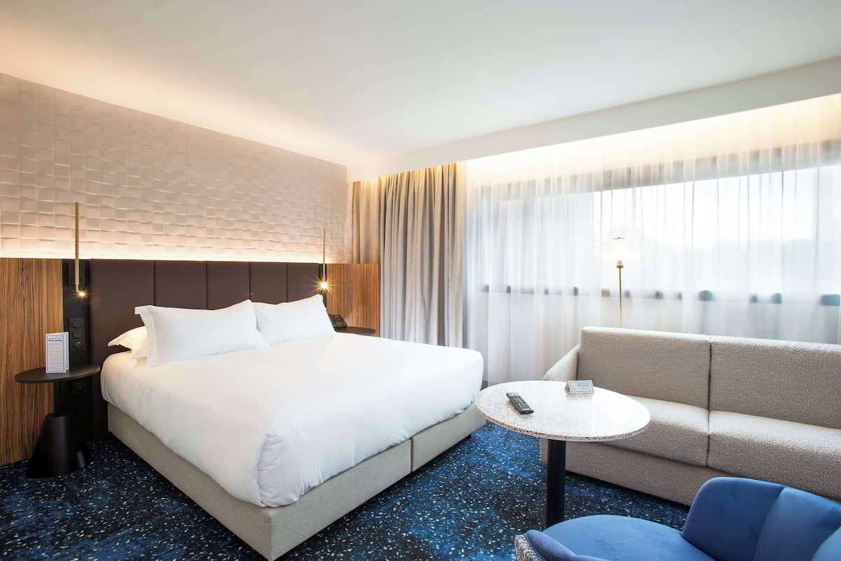 Une chambre d’hôtel avec un grand lit, un canapé, une petite table ronde et un panneau de commande fixé au mur.