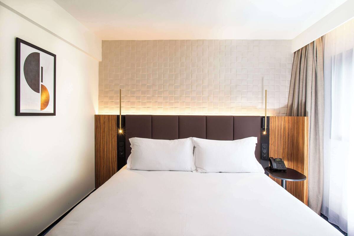 Une chambre d’hôtel avec un grand lit, une table de chevet avec un téléphone, une lampe fixée au mur et un tableau encadré.