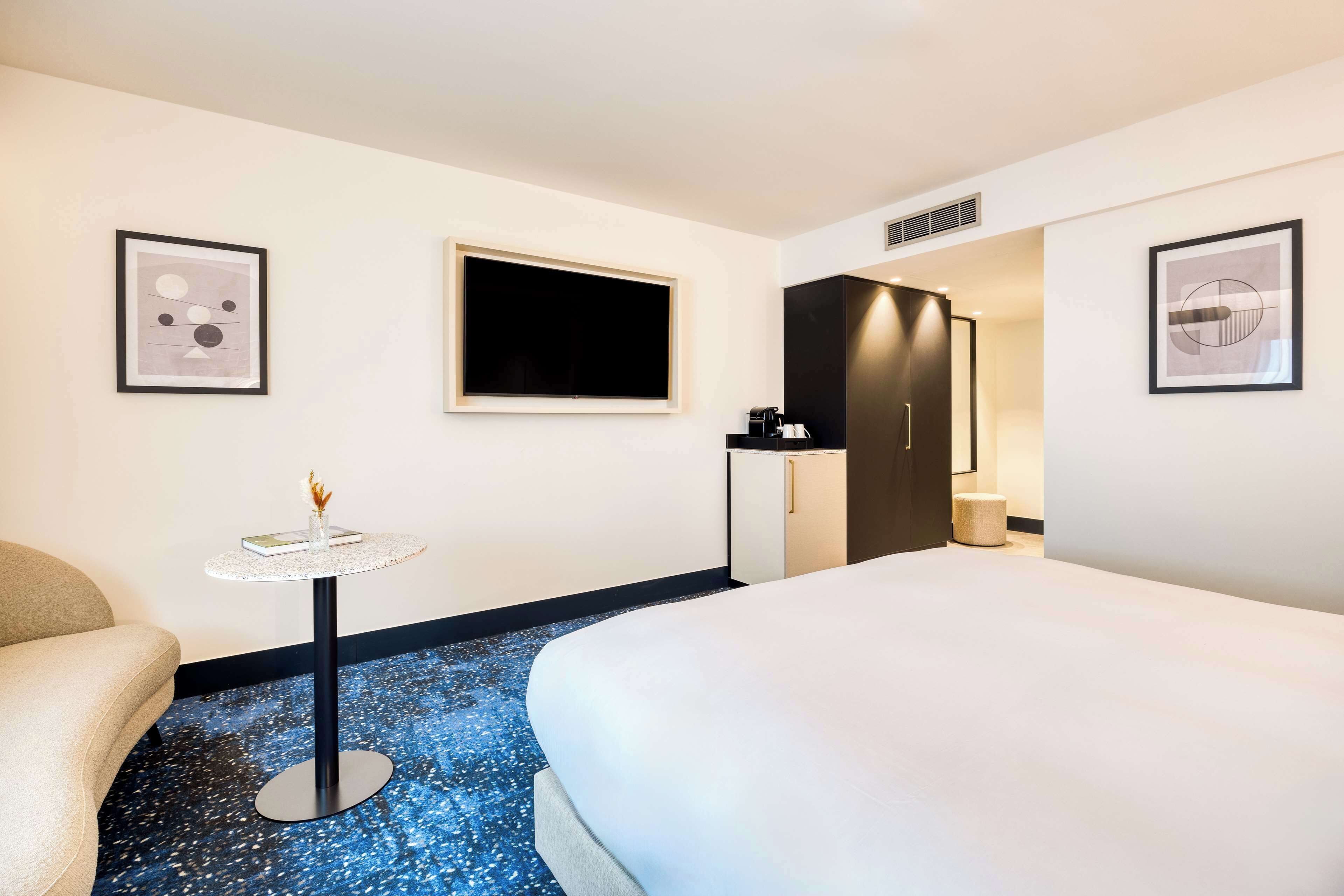A hotel room with a bed, a small round table with a vase, a TV, a wardrobe, and a framed picture on the wall.