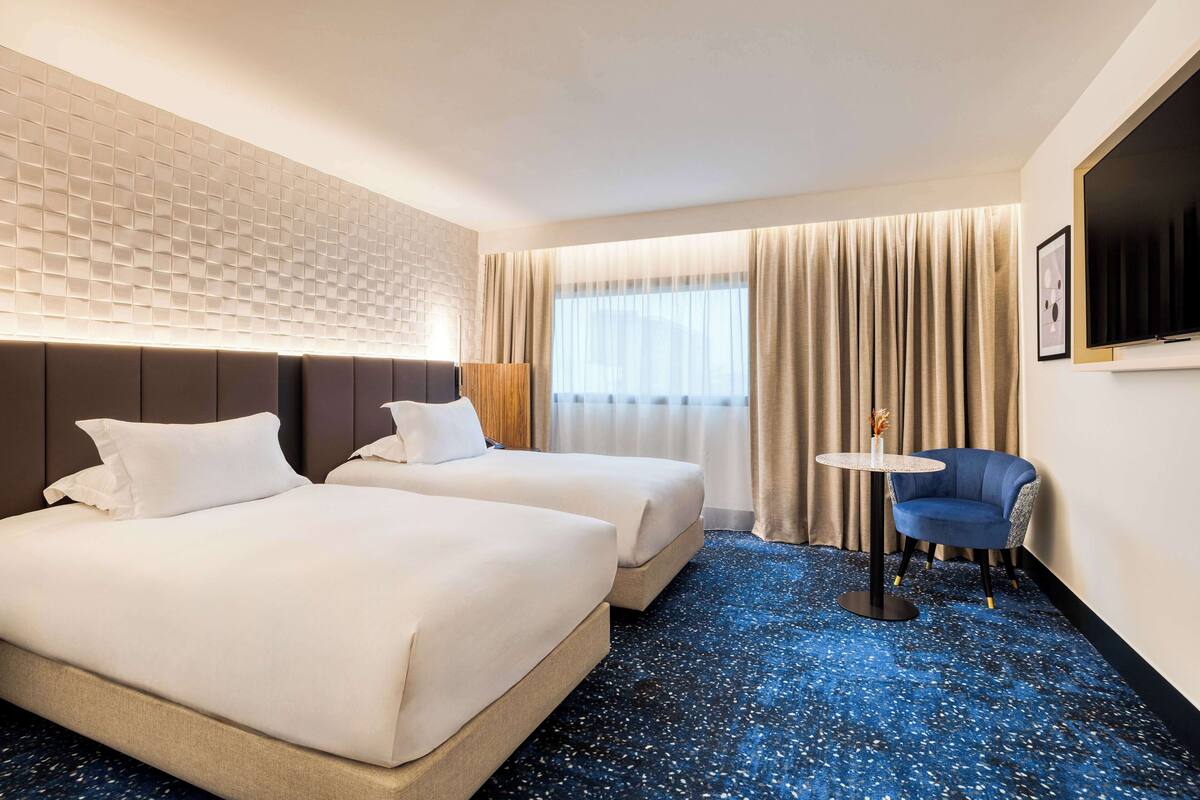 Une chambre d’hôtel avec deux lits, un fauteuil bleu, une petite table ronde, un téléviseur fixé au mur et une fenêtre avec des rideaux.
