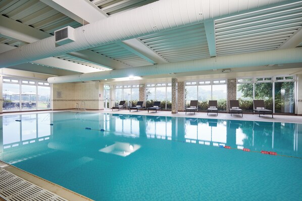 Una piscina interior con agua cristalina, sillas de descanso y grandes ventanales.