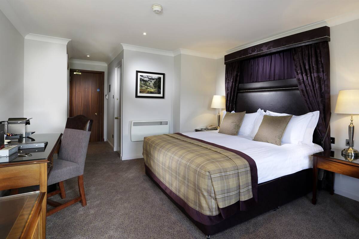 A hotel room with a bed, two bedside tables with lamps, a desk with a chair, a radiator, and a framed picture on the wall.