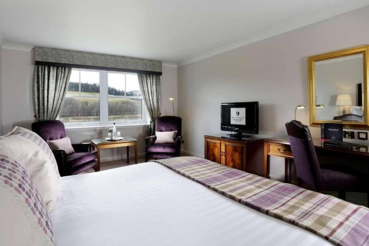 A hotel room with a large bed, two armchairs, a desk with a computer, a television, and a window with curtains.