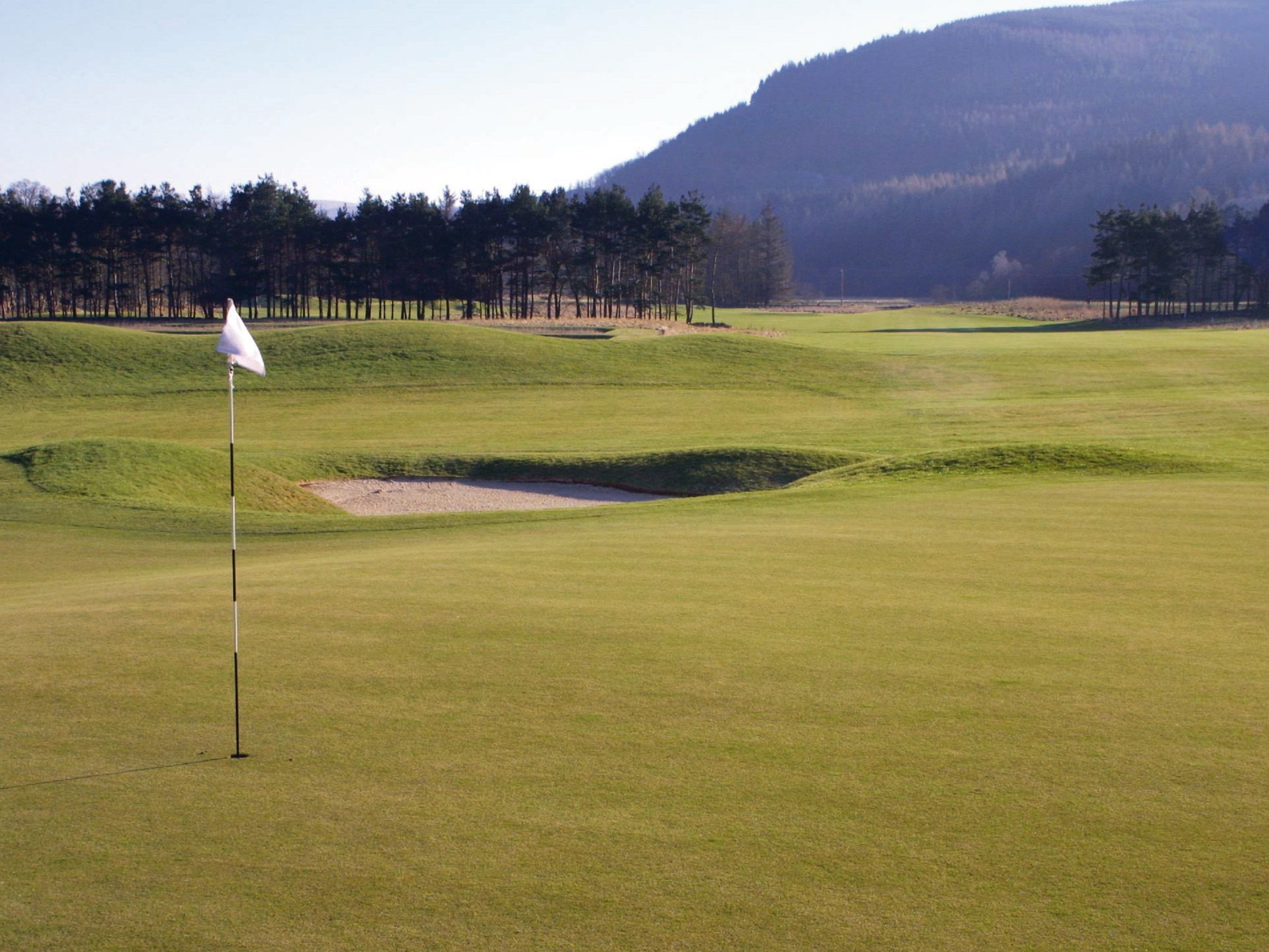 A golf course with a flagstick, sand bunkers, and a forested hill in the background.