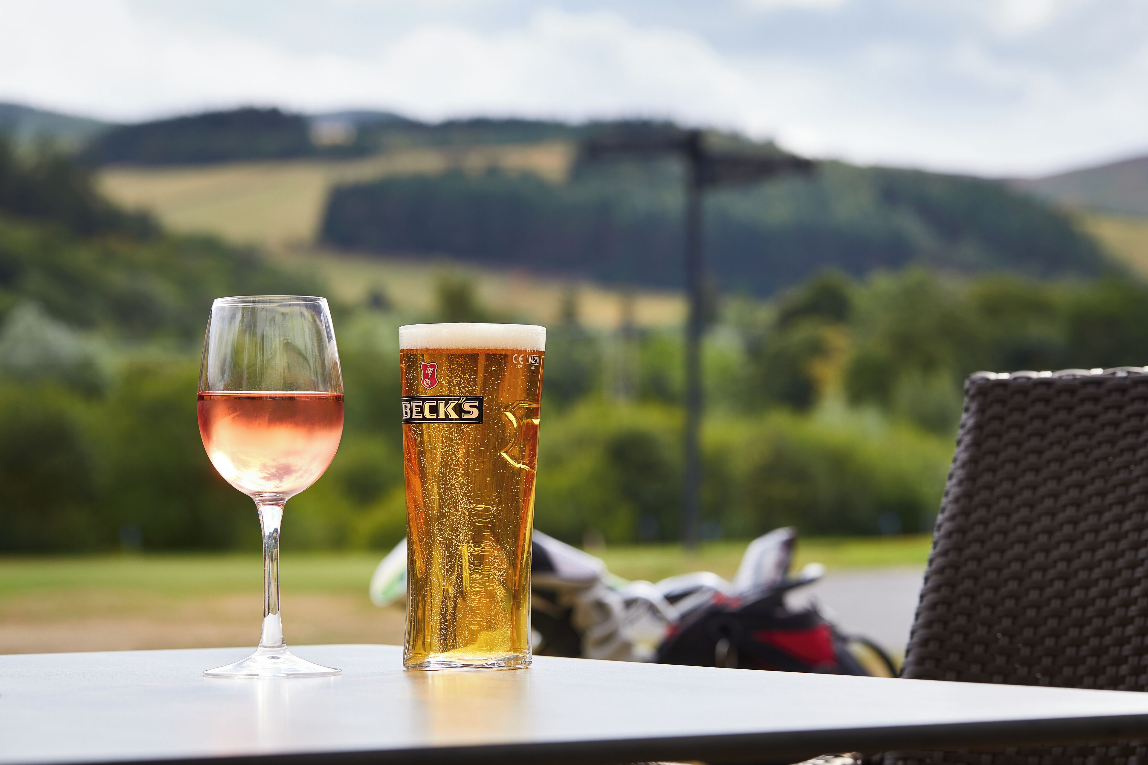 A table with a glass of beer and a tall glass of beer with a label, set against a scenic outdoor background.
