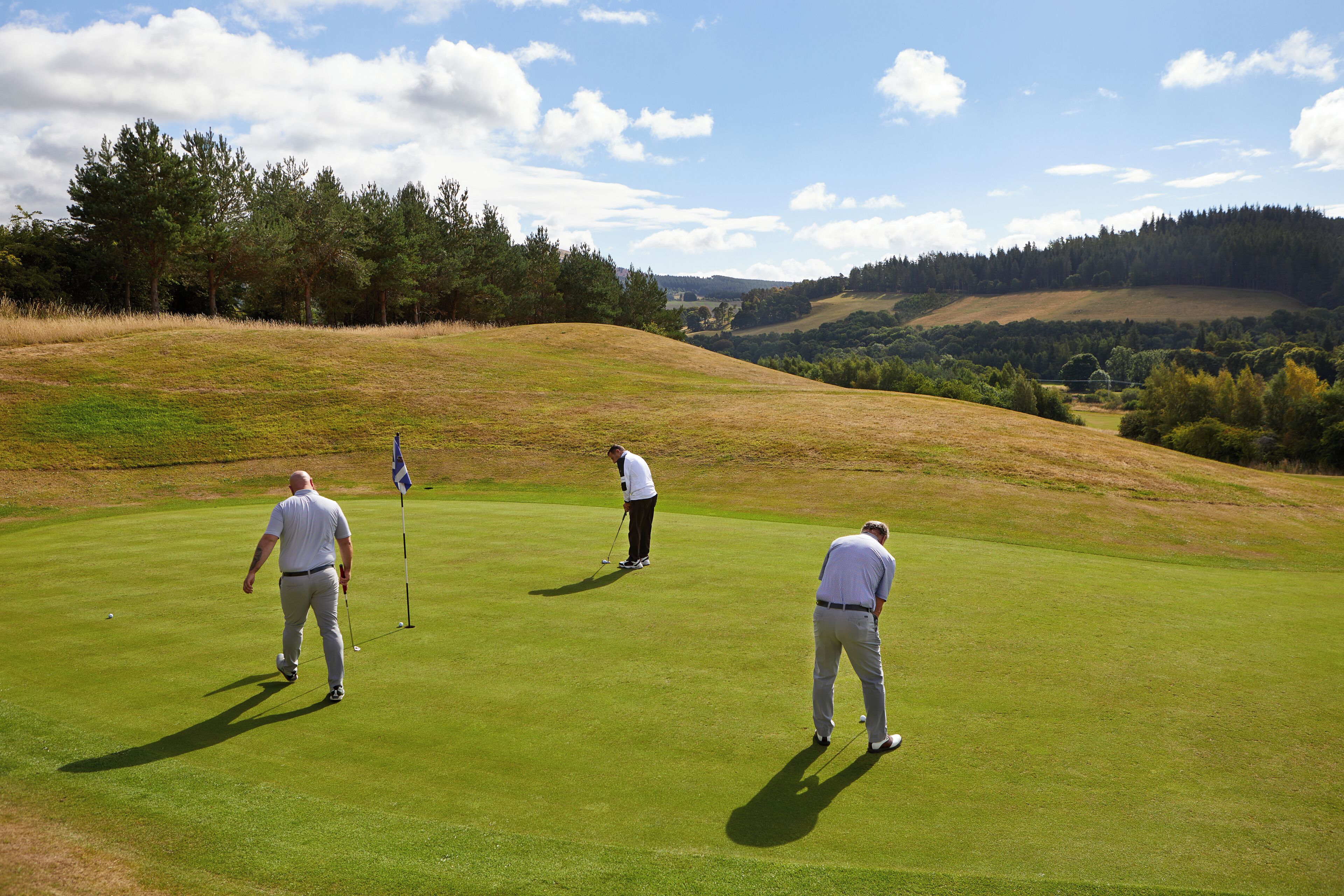Un campo de golf con tres jugadores en el green, rodeado de colinas ondulantes y árboles.
