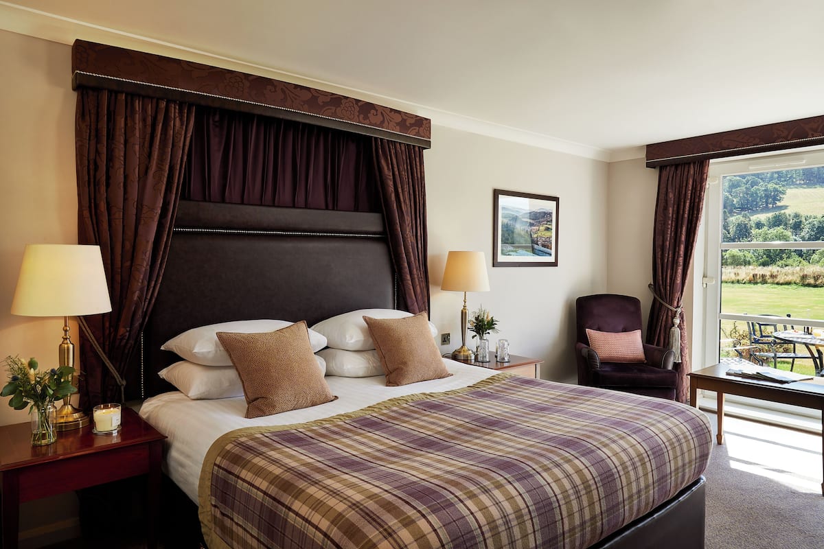 A hotel room with a large bed, two bedside tables with lamps, a chair, and a view of a green landscape through the window.