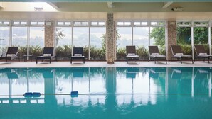 An indoor swimming pool with clear blue water, surrounded by lounge chairs and large windows offering views of greenery. - Macdonald Cardrona Hotel, Golf & Spa (Peebles)
