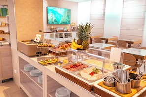 Zona de buffet con diversas opciones de comida, un televisor en la pared y un área de estar con mesas y sillas.