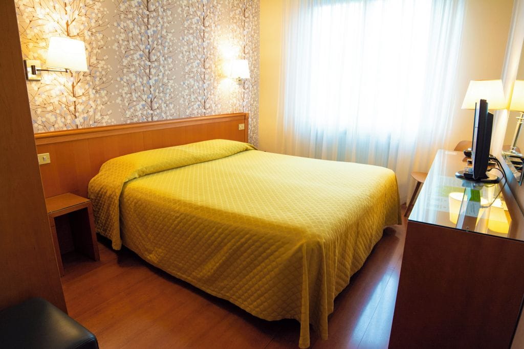 A hotel room with a bed, bedside table, lamp, and a wall with floral wallpaper.