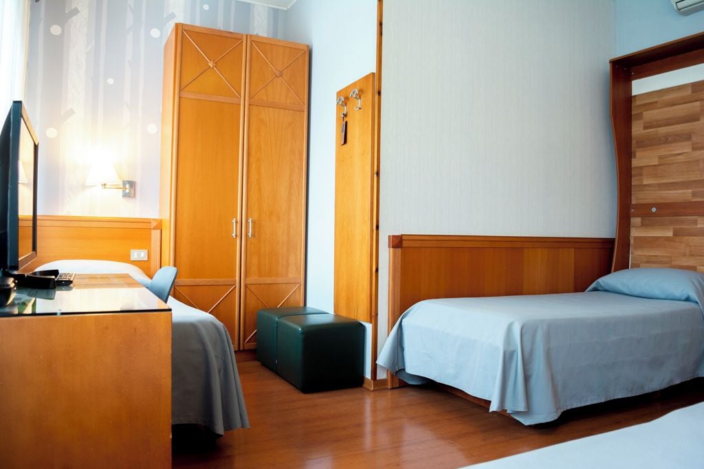 Une chambre d’hôtel avec deux lits, un bureau, une armoire et un petit pouf.