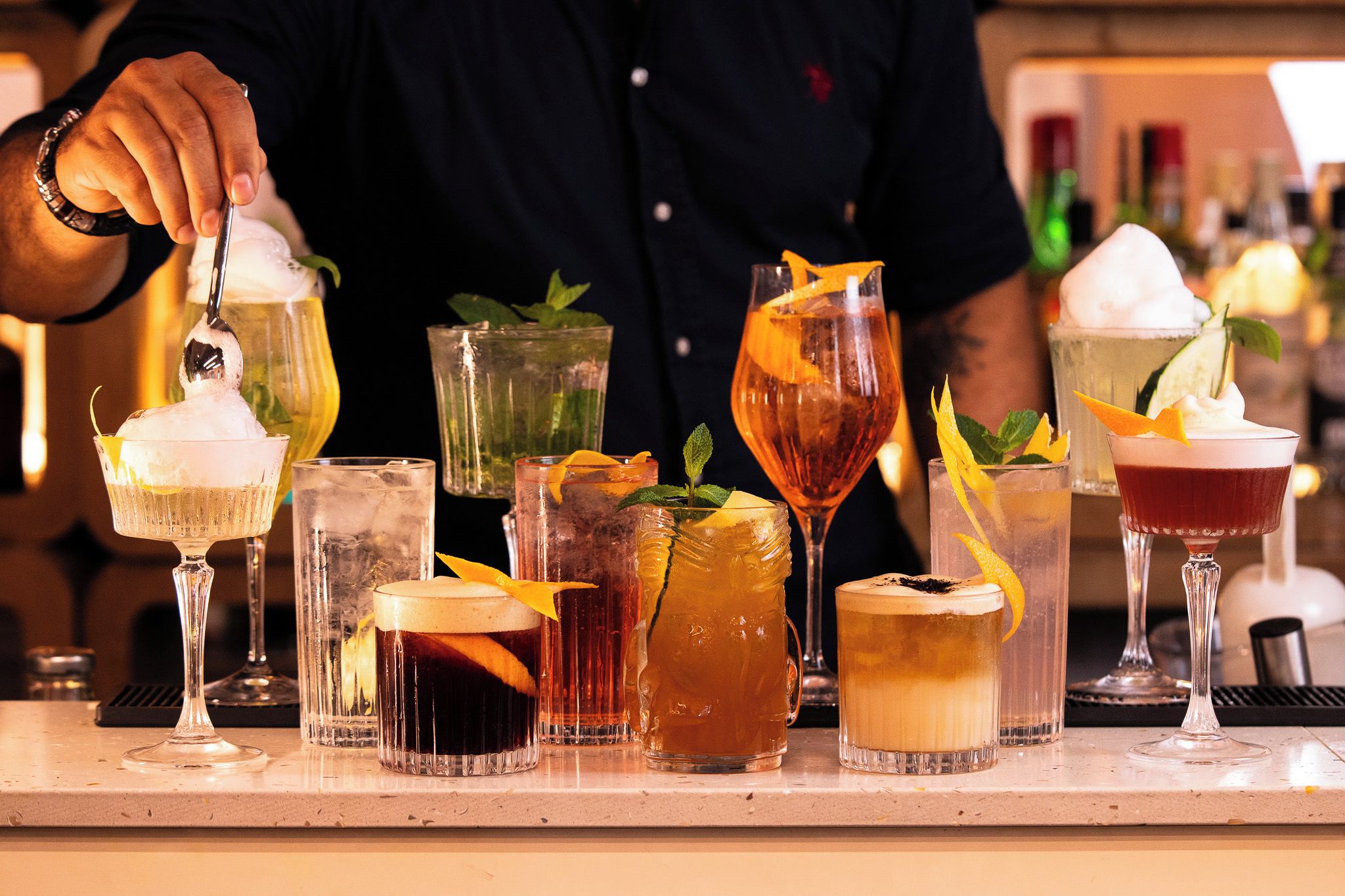 Un barman prépare des boissons derrière un comptoir, avec différents types de verres à cocktail et une carafe.