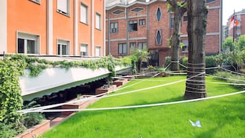Une zone herbeuse avec un arbre entouré de cordes, un bâtiment blanc doté d’un toit en pente et, au fond, un bâtiment en briques avec de nombreuses fenêtres et des balcons.
