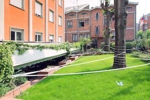 Un área con césped, un árbol envuelto en cuerda, un edificio blanco con techo inclinado y un edificio de ladrillo con varias ventanas y balcones al fondo.