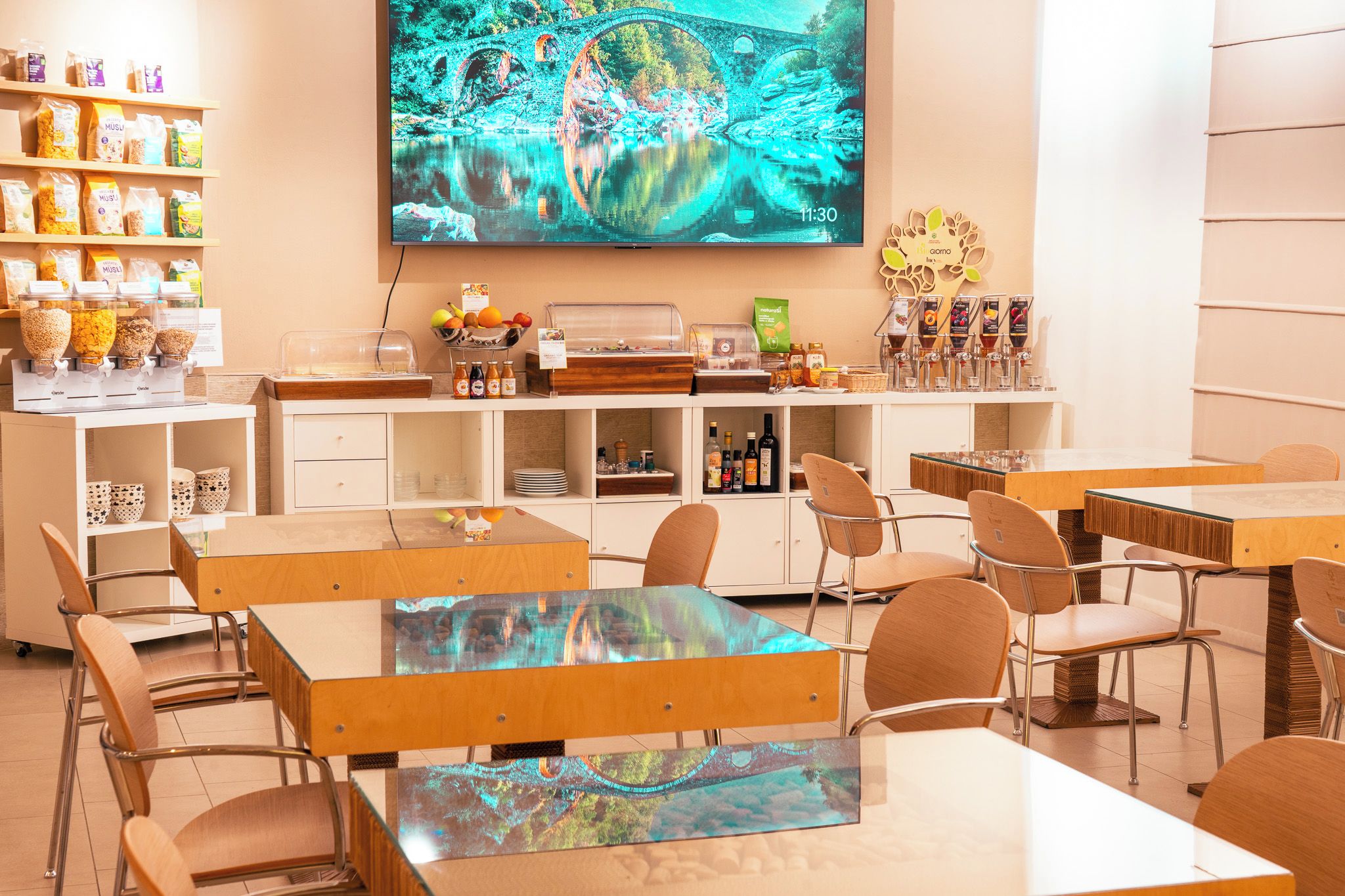 Un espace repas moderne avec des tables et des chaises en bois, un buffet proposant divers plats, et un grand écran diffusant une image panoramique.