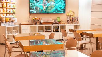 Un espace repas moderne avec des tables et des chaises en bois, un buffet proposant divers plats, et un grand écran diffusant une image panoramique.