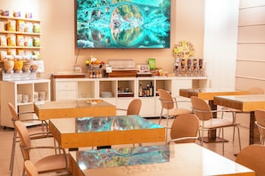 A modern dining area with wooden tables and chairs, a buffet with various food items, and a large screen displaying a scenic image. - eco Hotel Milano & BioRiso Restaurant (Milan)