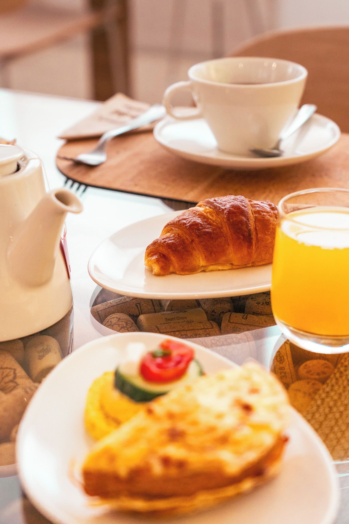 A breakfast setting with a croissant, a cup of coffee, a teapot, a glass of orange juice, and a plate with a slice of toast topped with a tomato and a slice of cheese.
