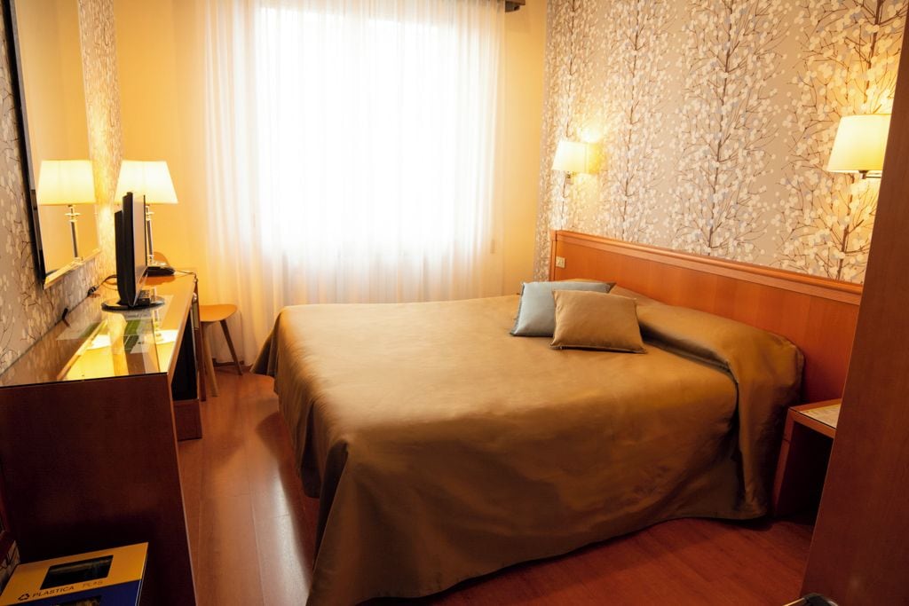 Quarto de hotel com cama, mesas de cabeceira, luminárias e uma janela com cortinas.