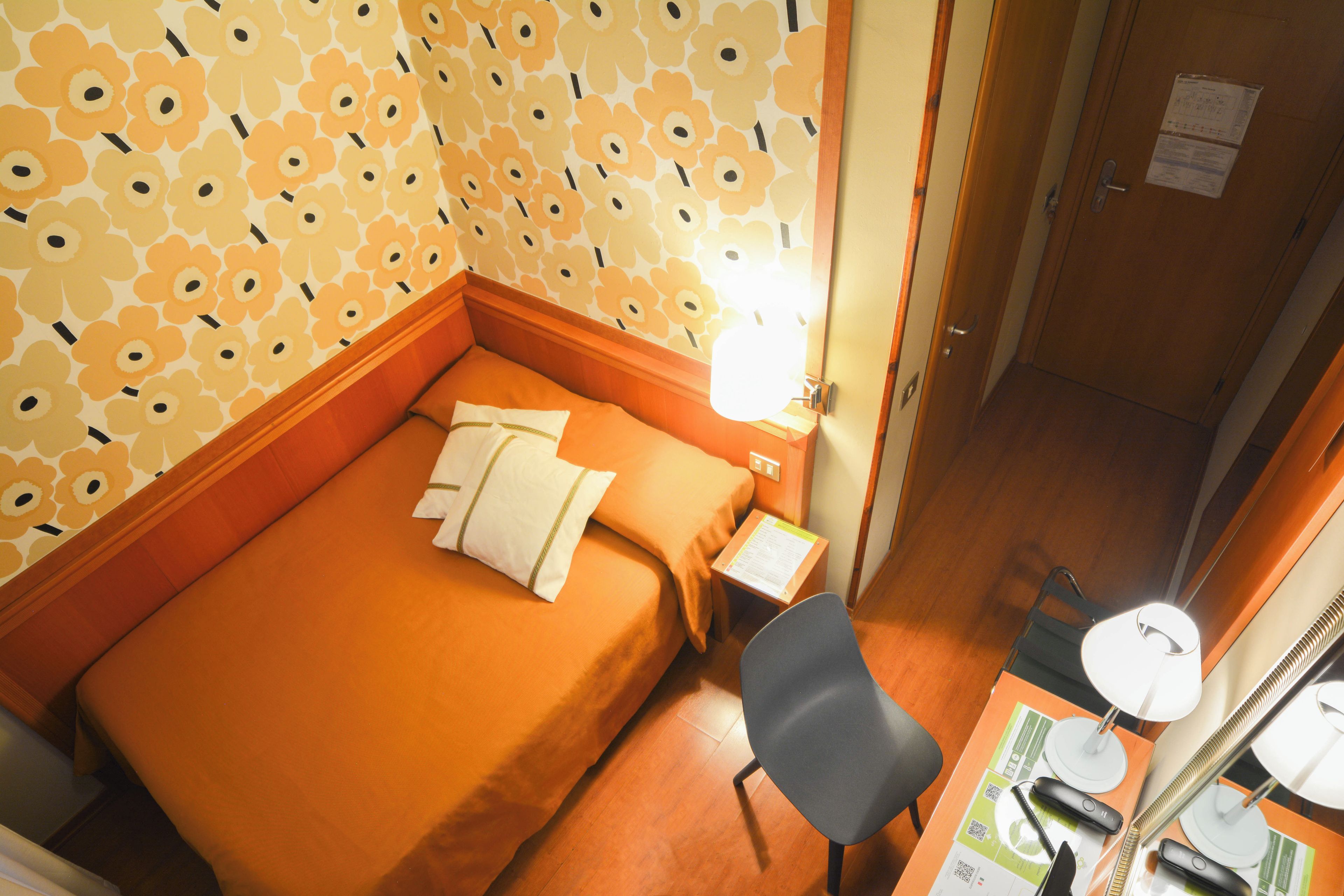 Une chambre d’hôtel avec un lit, un bureau et une chaise, une lampe et une porte sur laquelle est affiché un avis.