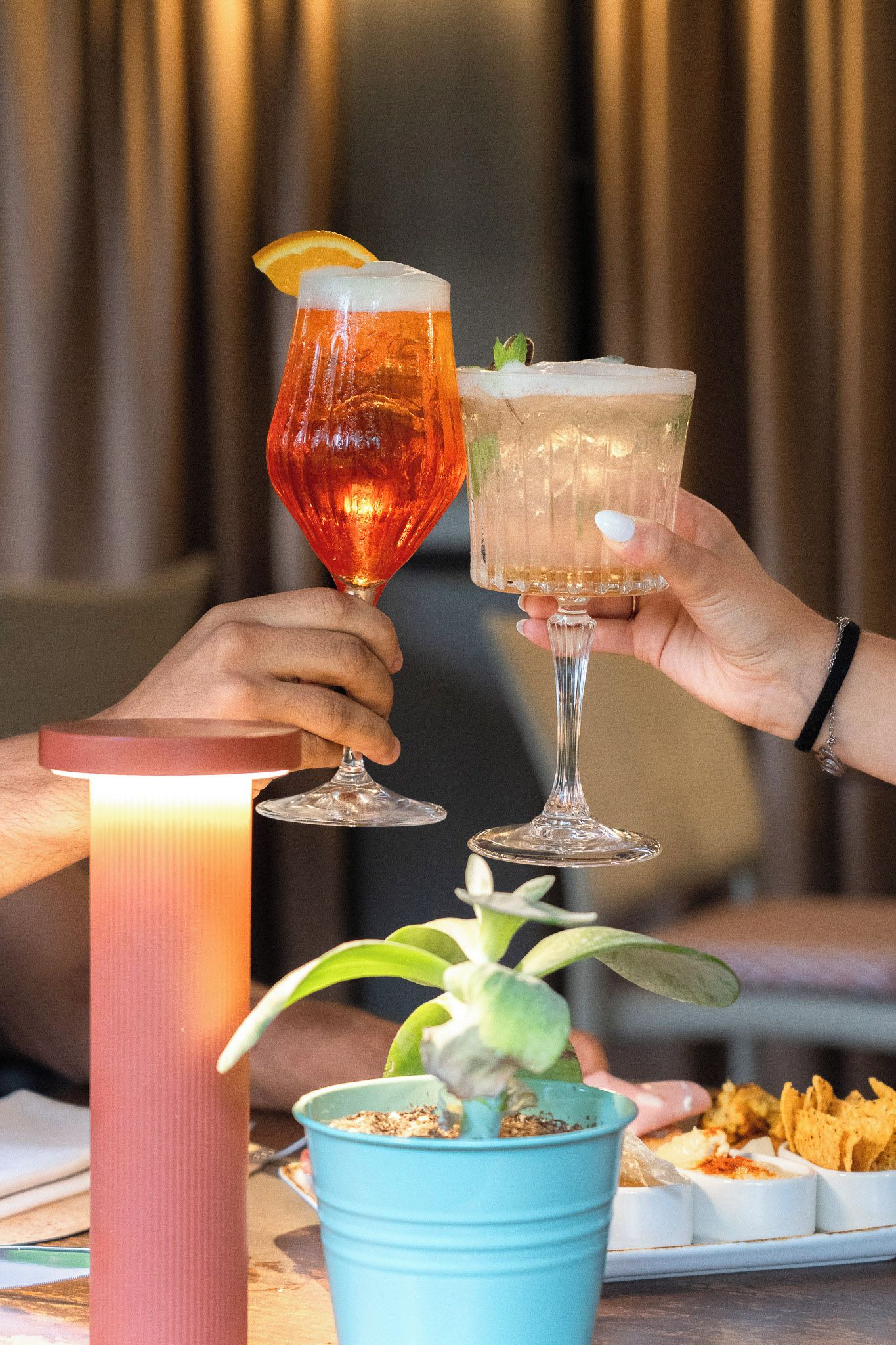 Deux personnes tenant des verres remplis de boissons, une plante et une assiette de nourriture sur une table.