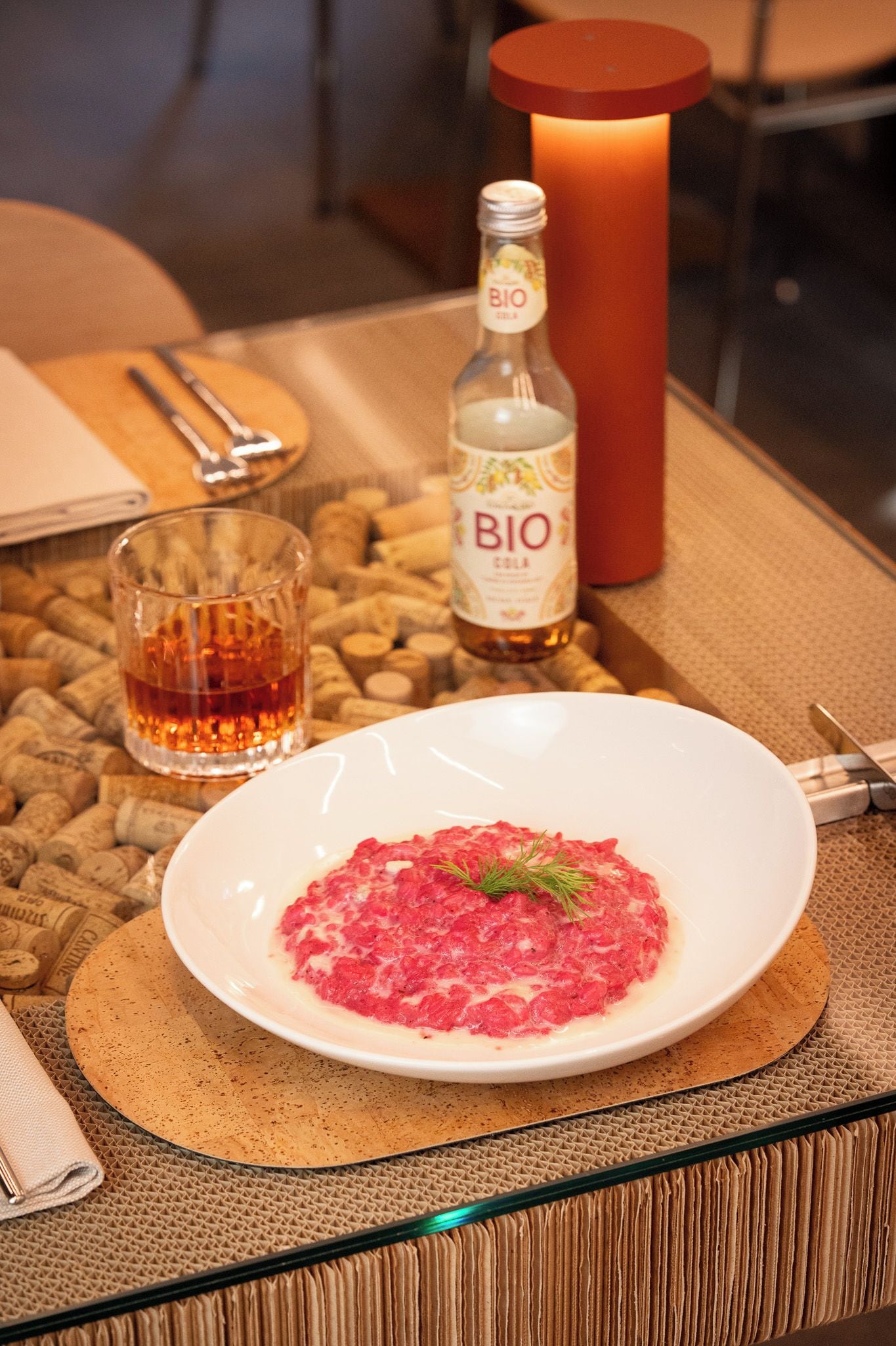 Un piatto di cibo, una bottiglia di BIO cola, un bicchiere di liquido ambrato e una candela su un tavolo con dei tappi di sughero.