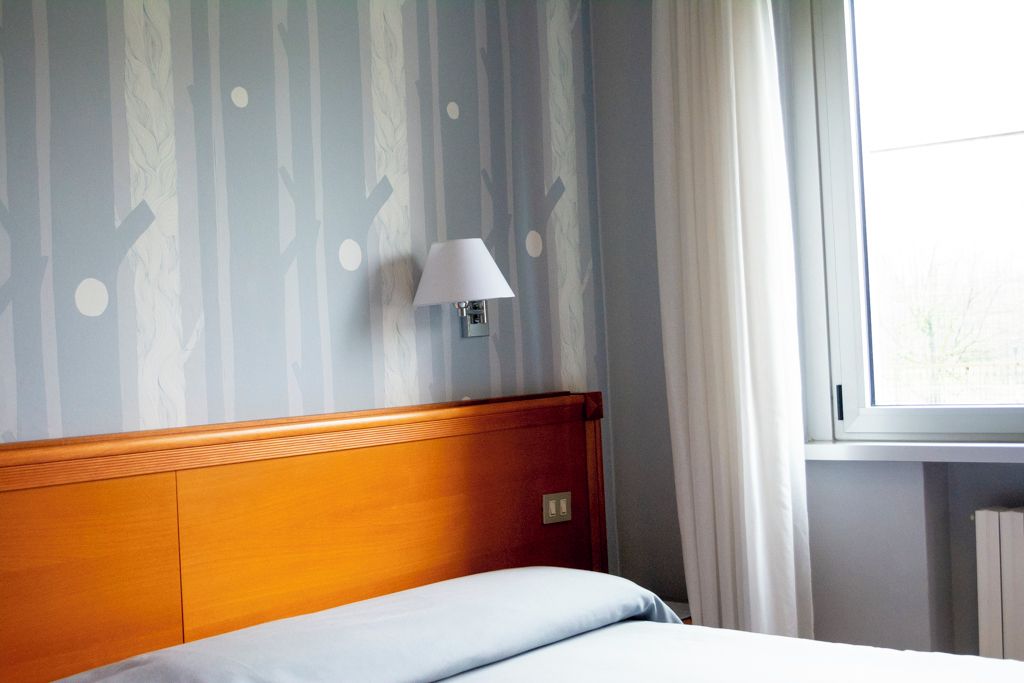 Ett hotellrum med en sänggavel i trä, en säng med ett vitt lakan, en vägglampa och ett fönster med gardiner.