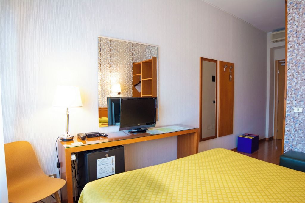 A hotel room with a bed, a desk with a television, a mirror, a wardrobe, and a small seating area.