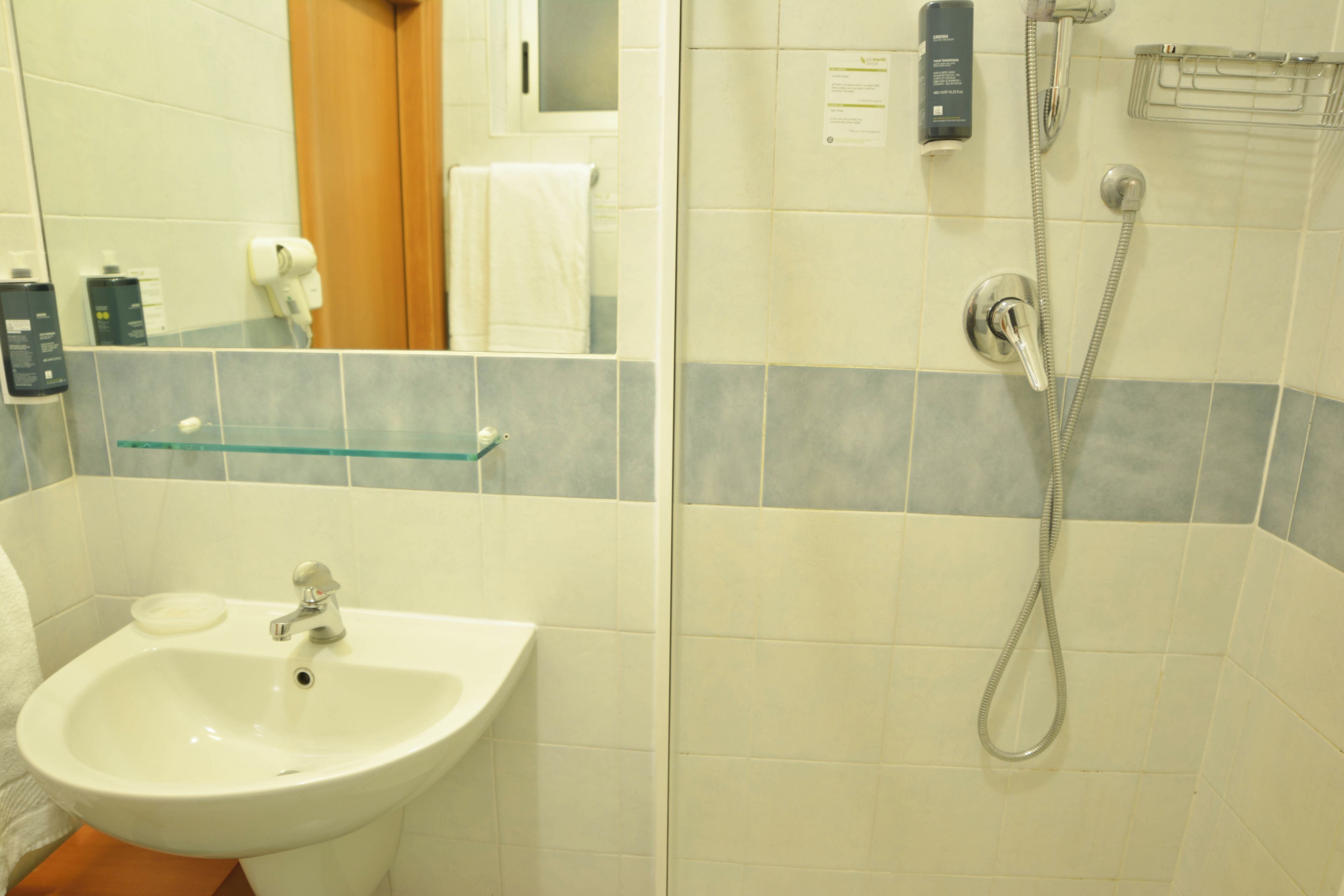 Une salle de bain avec un lavabo blanc, une étagère en verre, une douche équipée d’un pommeau de douche à main et un porte-serviettes.