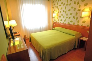 Una habitación de hotel con una cama, un escritorio con lámpara, una mesita de noche y una lámpara de pared.