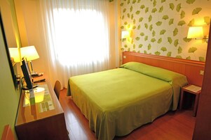 Una camera d'albergo con un letto, una scrivania con lampada, un comodino e una lampada a parete.