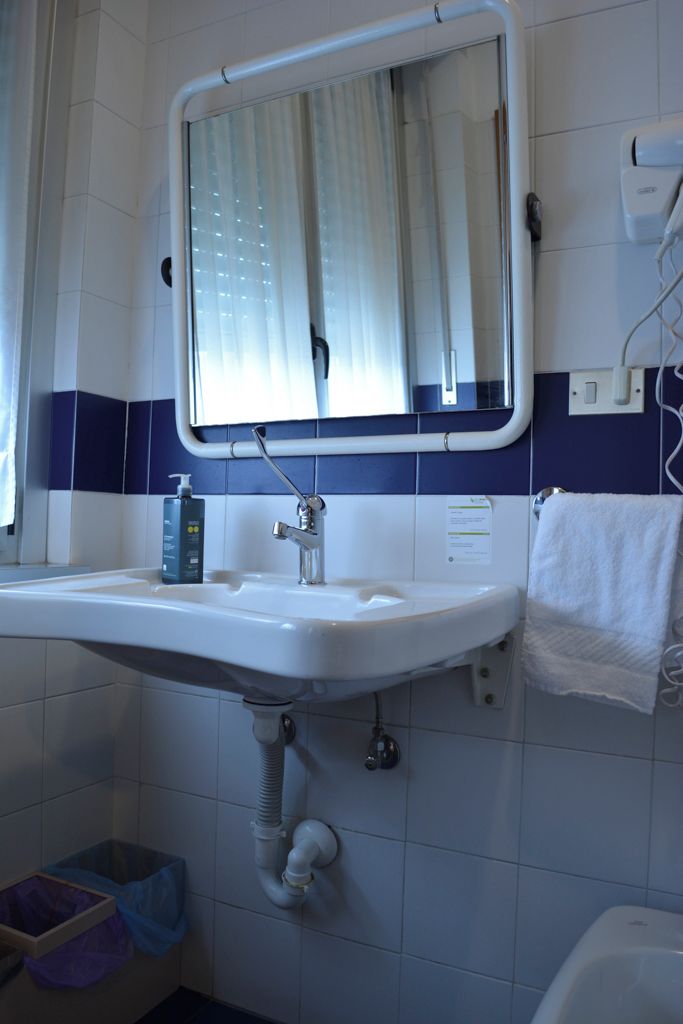 Une salle de bain avec un lavabo blanc, un miroir avec une fenêtre, un porte-serviettes et une poubelle.