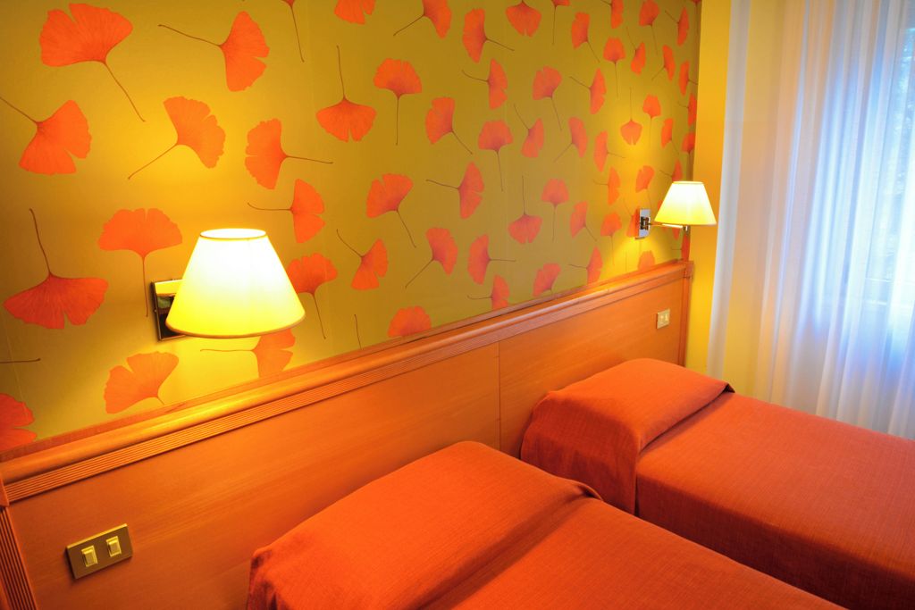 Ein Hotelzimmer mit zwei Betten, einer Wand mit Blattmuster, einer Lampe und einem Fenster mit Vorhängen.