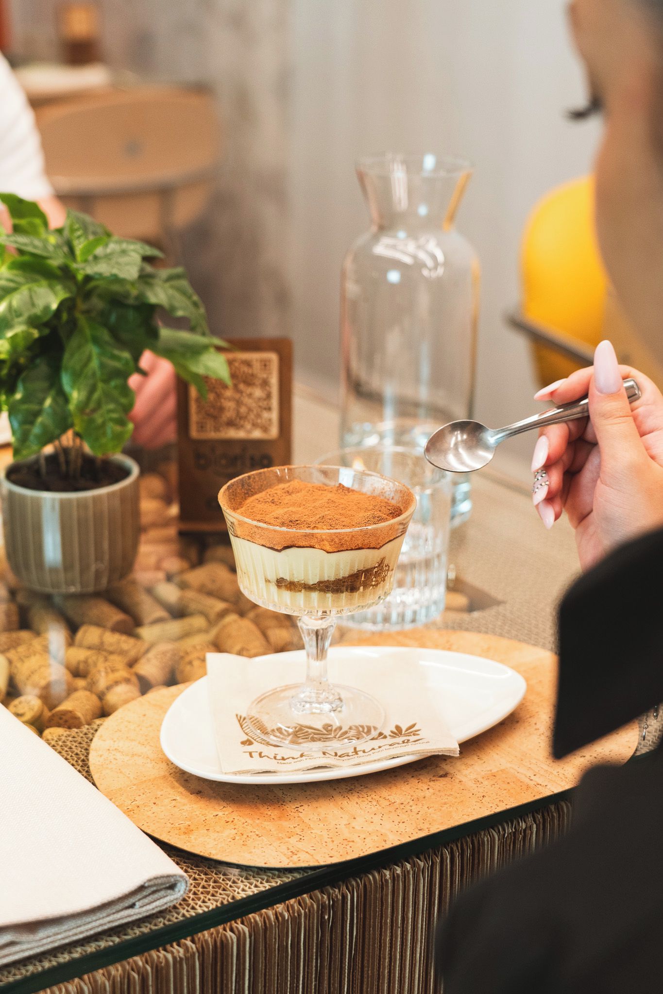 Postre en un bol de vidrio sobre un plato con cuchara, planta en maceta, botella y tapones sobre una mesa.