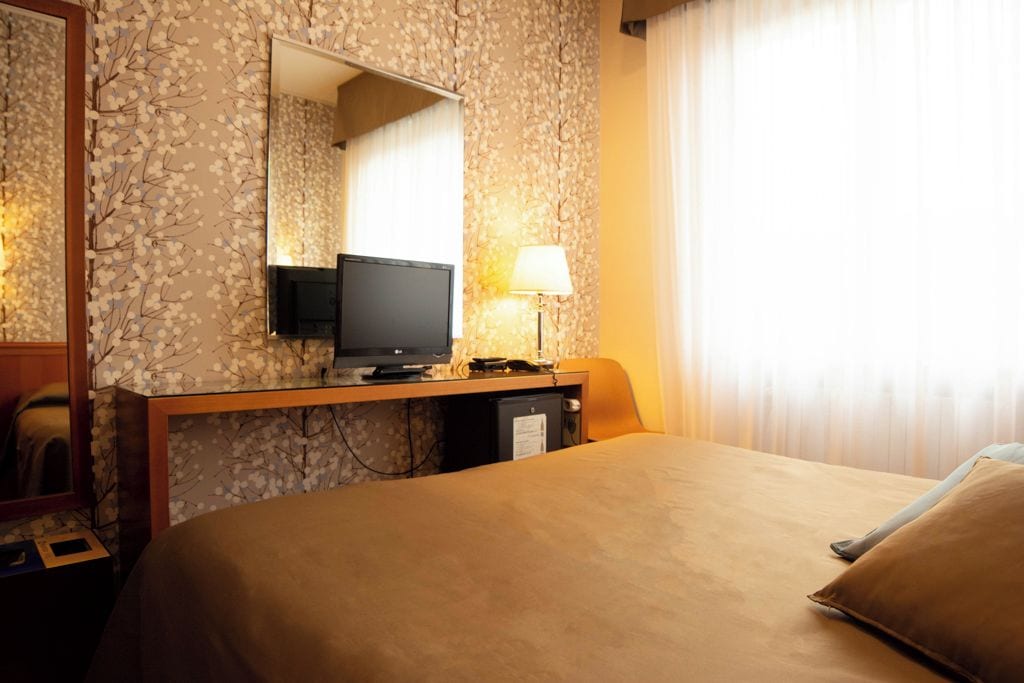 A hotel room with a bed, a desk with a television, a lamp, and a mirror.