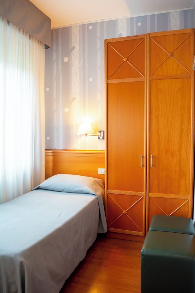 Une chambre d’hôtel avec un lit, une armoire en bois, une table de chevet avec une lampe et un banc vert.