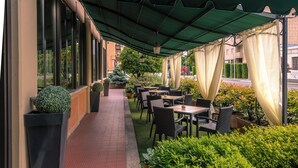 Un café al aire libre con mesas y sillas, plantas en macetas y un porche cubierto.