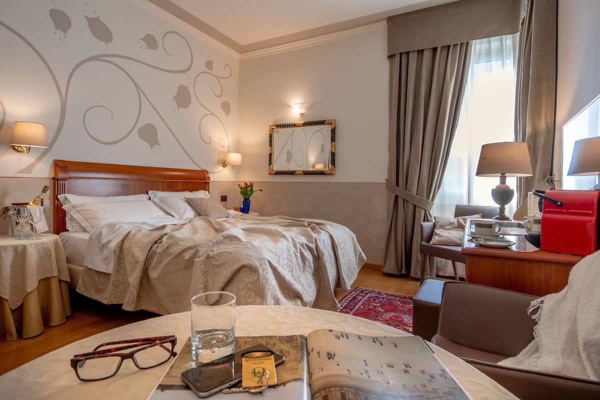 A hotel room with a large bed, bedside tables with lamps, a desk with a red printer, and a window with curtains.