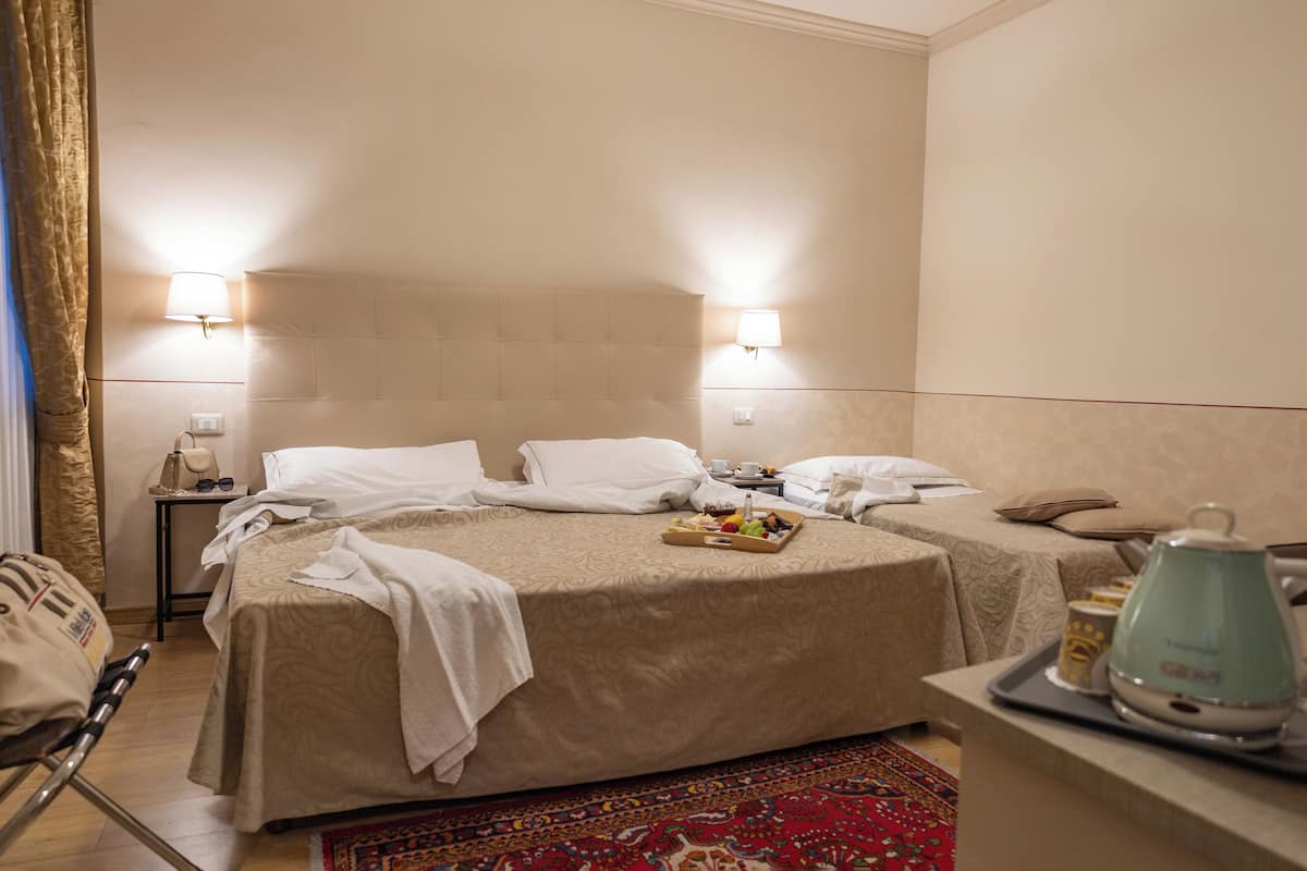 A hotel room with two beds, a small table with a kettle and cups, and a bedside table with a lamp and a handbag.