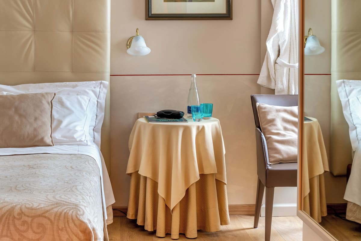 A hotel room with a bed, a small table with a lamp, a chair, a framed picture on the wall, and a robe hanging on a hook.