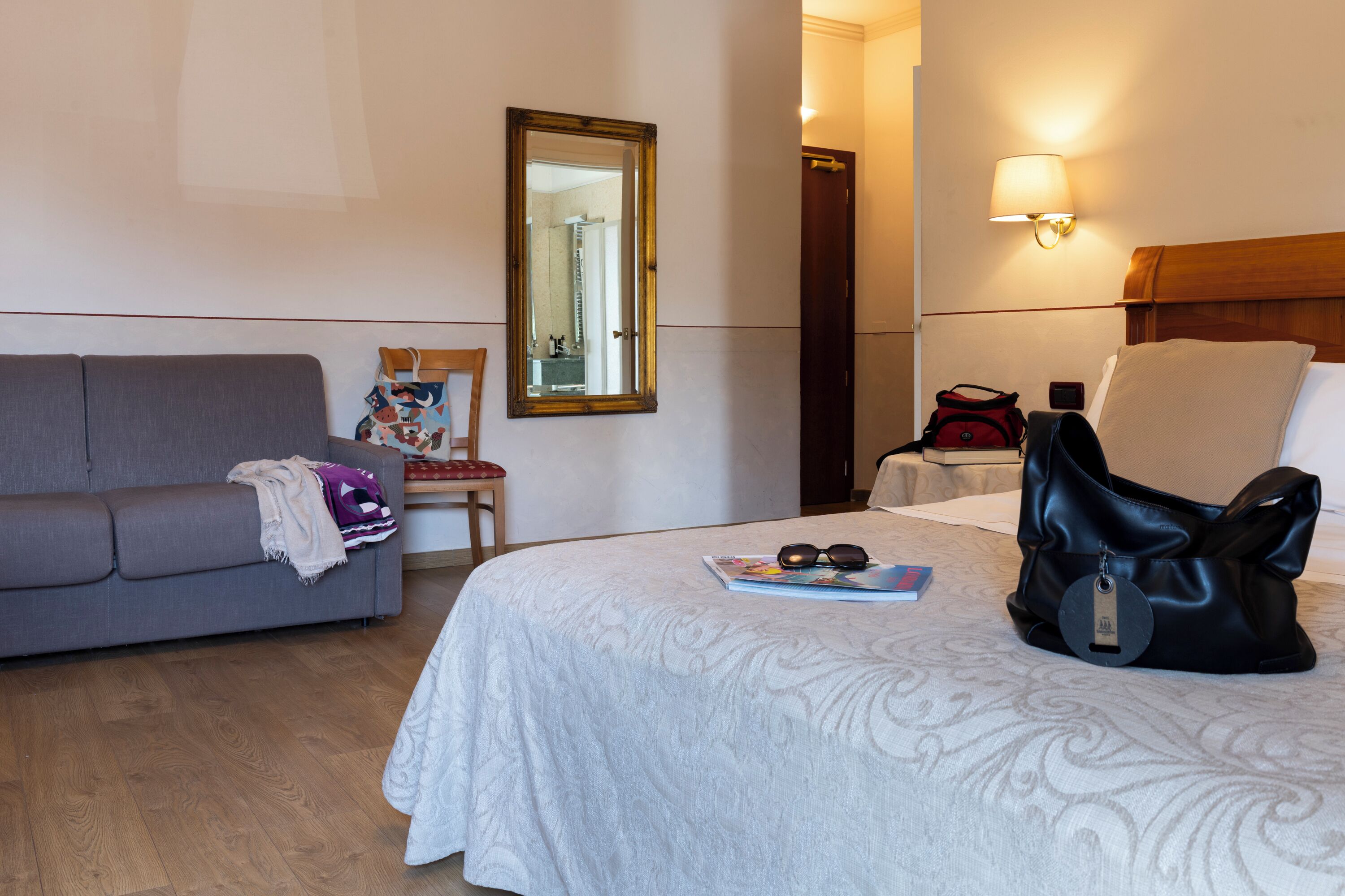 A hotel room with a bed, a gray sofa, a chair, a mirror, and a lamp.