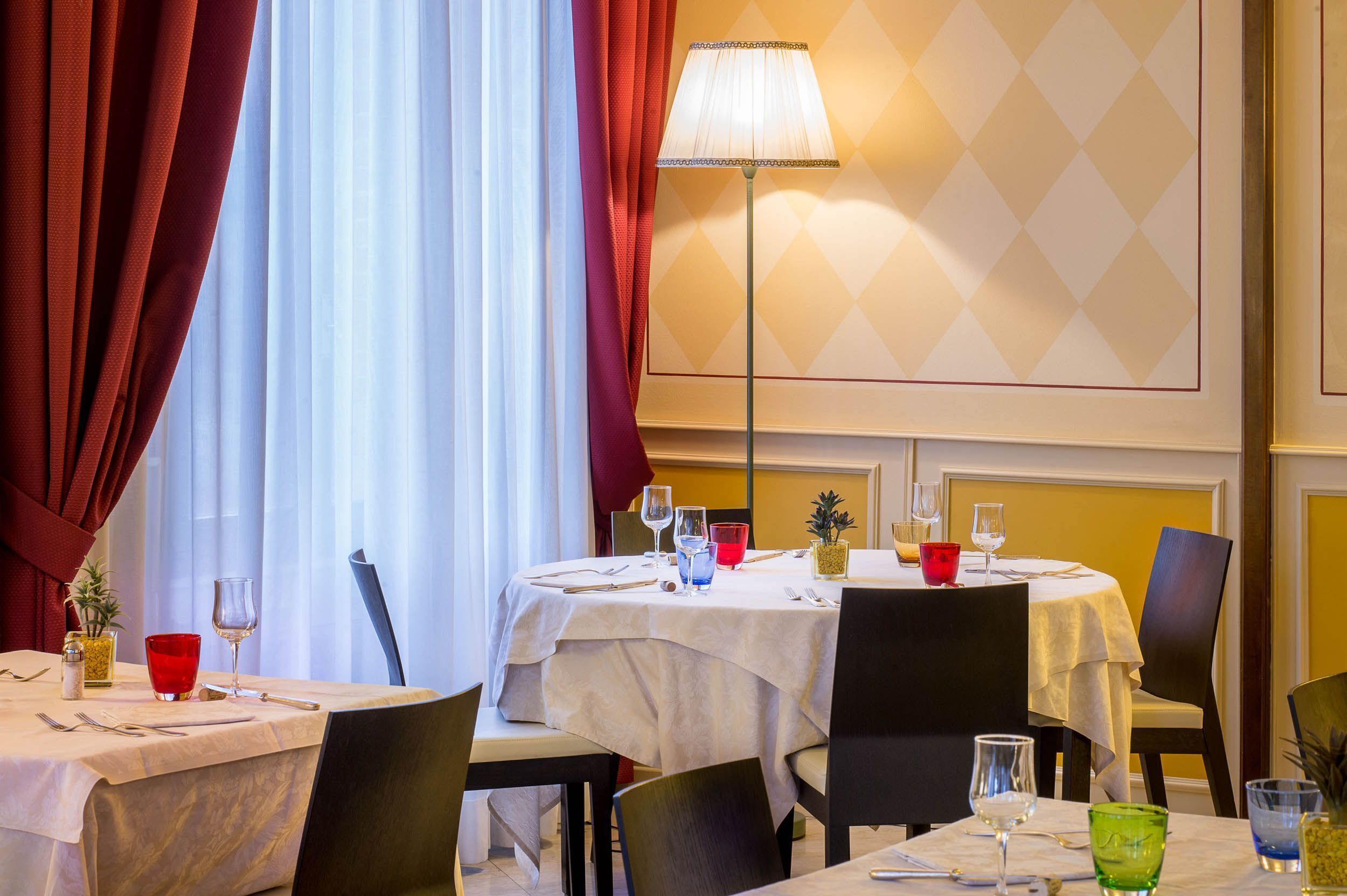Un espace repas avec des tables préparées pour deux personnes, agrémenté de nappes blanches, de verres rouges et verts, et d’un lampadaire.