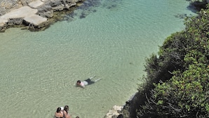 Una zona costera rocosa con agua cristalina, gente nadando y paseando por la orilla.