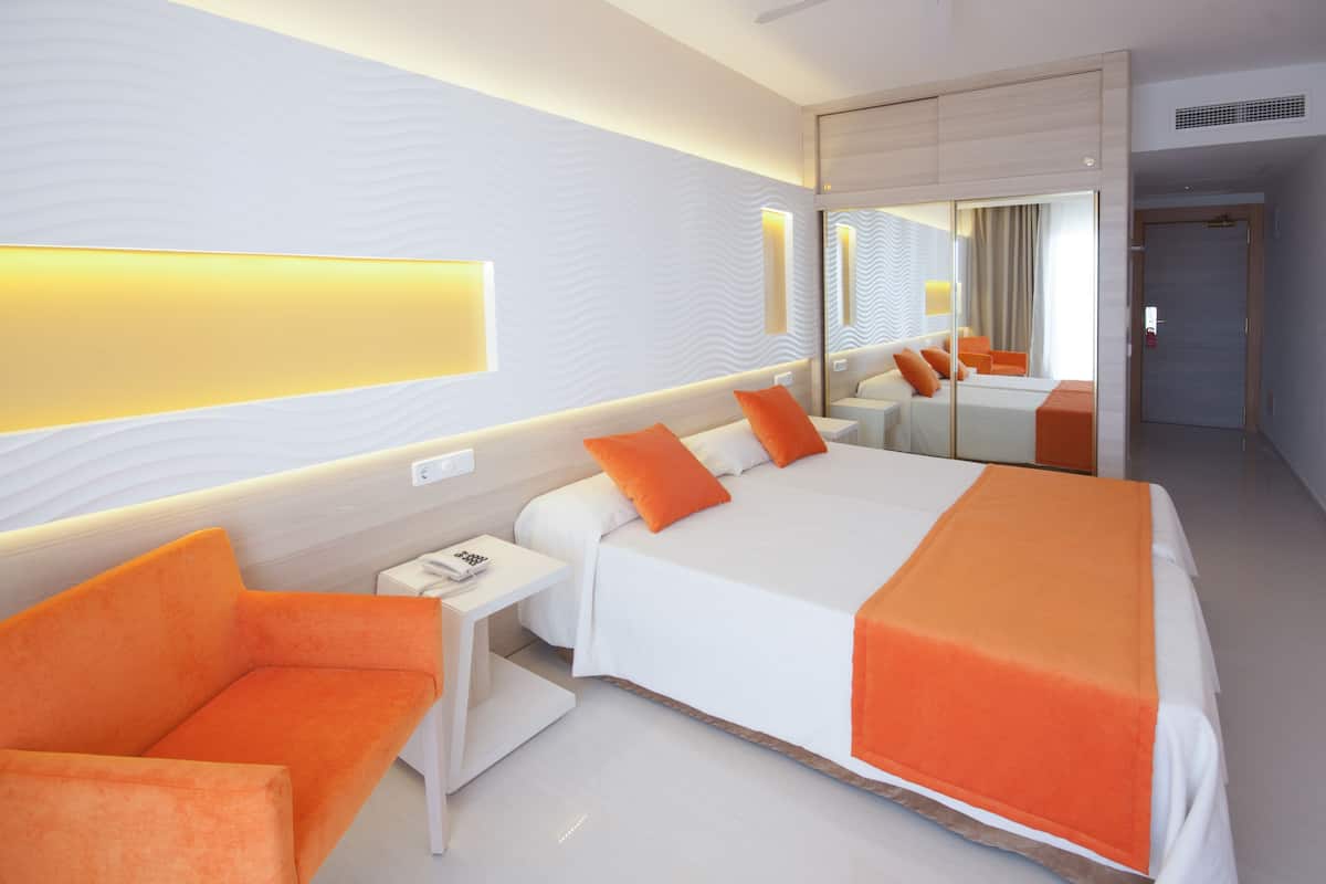 Ein modernes Hotelzimmer mit einem großen Bett, orangefarbenen Ziertelementen, einem Nachttisch mit Telefon und einem Spiegel, der den Raum reflektiert.