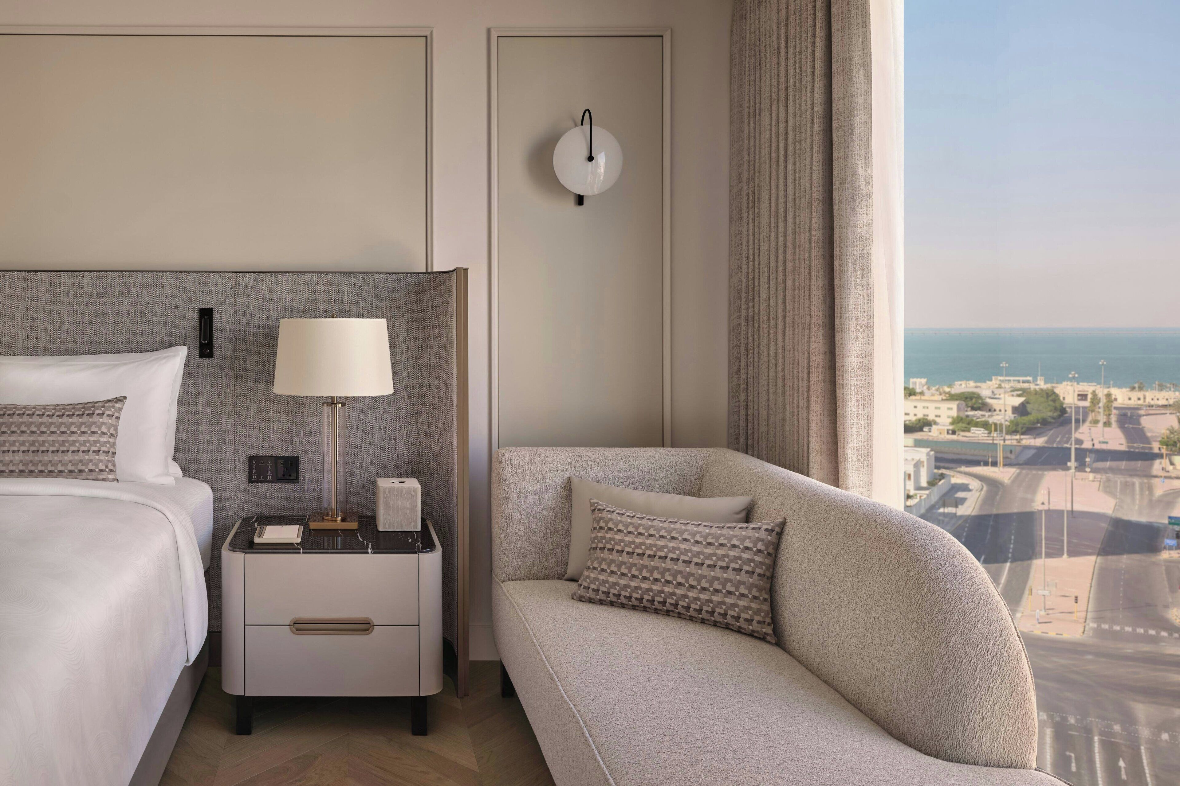 A hotel room with a bed, nightstand, lamp, and a sofa with a view of the city.